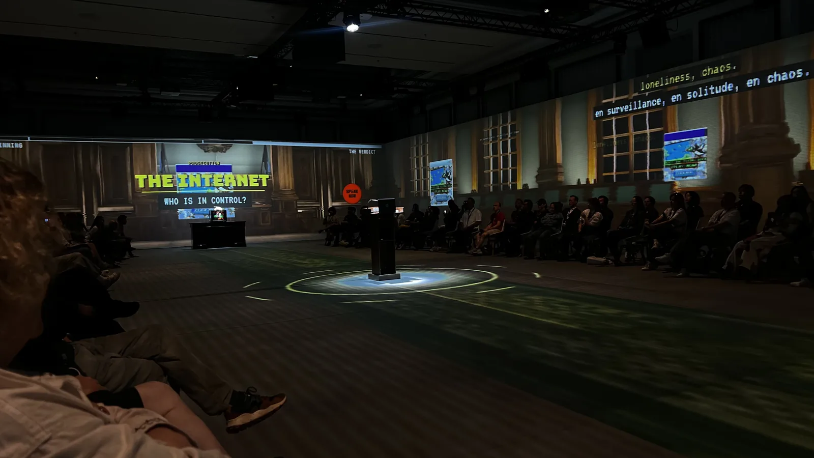 An audience surrounds the projected courtroom of GAWD v. The People, with a witness lectern in the centre.